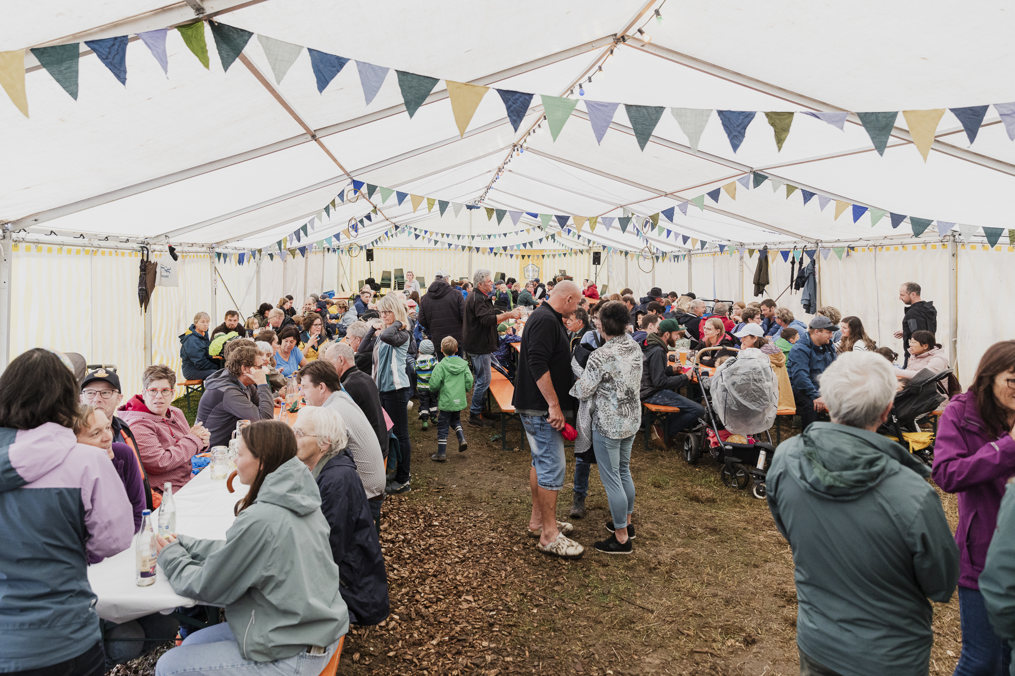 Besucherinnen und Besucher im Festzelt. Zu sehen sind viele Personen, die an Bierbänken sitzen und die Speisen genießen. Geschmückt ist das Zelt mit bunten Fähnchen. 